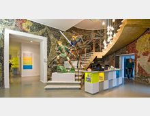 Foyer eines Museums mit einer geschwungenen Treppe, Mosaikwänden und einer Skulptur von zwei Fantasy-Figuren mit Schwertern. Im Vordergrund steht ein Empfangstresen und im Hintergrund führen Türen in weitere Ausstellungsräume.