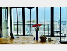 Ein modern eingerichteter Raum mit einem Holztisch, darauf eine Vase mit roten Blumen, eine Schale und ein Laptop. Durch die bodentiefen Glasfenster ist ein Balkon mit Sitzmöbeln sichtbar, im Hintergrund der Berliner Fernsehturm.