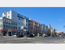 Straßenszene mit mehrstöckigen Wohn- und Geschäftsgebäuden, darunter ein modernes Gebäude mit weißem Putz und ein roter Backsteinbau. Im Vordergrund befinden sich Verkehrszeichen, eine Straße mit Autos und eine Fußgängerampel; blauer Himmel im Hintergrund.