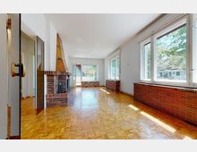Großzügiger Wohnraum mit hellem Parkettboden, bodentiefen Fenstern auf der rechten Seite und einem Kamin mit Backsteinverkleidung auf der linken Seite. Der Raum ist lichtdurchflutet und bietet Blick auf einen Garten durch die Fenster.