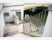 Abgang zu öffentlichen Toiletten mit den Schildern „Herrendamen“ und „Damen-WC“, gelegen unter einer Brücke neben einem Gewässer. Links und rechts der Treppe befinden sich graue Geländer, darunter eine grüne Tür.