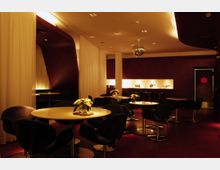 Ein elegantes, dimm beleuchtetes Café oder Lounge mit runden Tischen, schwarzen Sesseln und weißer Tischdekoration. Im Hintergrund sind eine Bar mit hohen Hockern, beleuchtete Nischen mit Dekorationsobjekten und weiße Vorhänge zu sehen; die Decke ist mit integrierter Beleuchtung und einer kleinen Discokugel gestaltet.