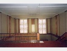 Innenansicht eines historischen Treppenhauses mit einer Holzbalustrade, großen Fenstern mit mehrfach unterteilten Scheiben und einem hellen Deckenbereich mit schlichten Verzierungen. Die Umgebung wirkt geradlinig und dezent gestaltet.