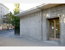 Außenansicht eines modernen Gebäudes mit einer Fassade aus großen grauen Steinplatten und einem gläsernen Eingang mit Holzrahmen. Im Hintergrund sind eine städtische Straße mit parkenden Autos, Bäumen und historischen Gebäuden zu sehen.