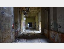 Ein verlassener, heruntergekommener Flur in einem Gebäude mit gelblichen Wänden, die stark mit Graffiti bedeckt sind. Der Boden ist beschädigt und mit Schutt übersät, die Decke zeigt sichtbare Verfallserscheinungen, und Licht dringt durch eine Türöffnung am Ende des Flurs.