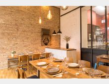 Gemütlicher Raum eines Restaurants mit Holztischen und gedeckten Plätzen, dekoriert mit Geschirr, Gläsern und Servietten. Die Wände bestehen aus Sichtziegeln und weiß gestrichenem Mauerwerk, ergänzt durch hängende Lampen und ein modernes Schild; eine Glaswand trennt die Küche vom Essbereich.