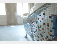 Nahaufnahme einer freistehenden Badewanne mit einer Außenseite, die mit bunten Mosaikfliesen in verschiedenen Farben verziert ist. Im Hintergrund ist ein heller, leerer Raum mit großen Fenstern und unverputzten Wänden erkennbar.