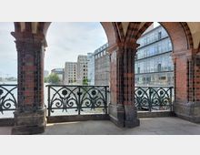 Blick durch die roten Backsteinbögen der Oberbaumbrücke in Berlin auf die umliegenden Gebäude am Spreeufer, darunter moderne Glasfassaden und historische Gebäudefassaden. Im Vordergrund ein kunstvoll gestaltetes Geländer mit floralen Motiven.