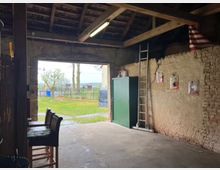 Innenansicht einer rustikalen Scheune mit Stein- und Putzwänden, einer Holzdecke und freiliegenden Balken. An der rechten Wand sind Poster angebracht, eine Leiter lehnt daran, und neben ihr steht ein grüner Metallschrank. Der offene Scheuneneingang gewährt den Blick auf einen Garten mit Bäumen und einem Zaun.