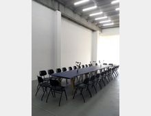 Ein minimalistischer Raum mit Betonwänden und einer sichtbaren Betondecke, beleuchtet durch Deckenleuchten. In der Mitte steht ein langer Tisch mit dunkler Tischplatte, umgeben von schwarzen Stühlen, auf dem einzelne Vasen mit roten Blumen stehen.