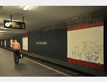 U-Bahnhof mit der Beschilderung „Kaiserin-Augusta-Str.“, grauen Betonwänden und roten Wandpaneelen. Ein Mann mit Tasche läuft auf dem Bahnsteig, und ein Zugzielanzeiger sowie mehrere Wände mit Graffiti sind sichtbar.