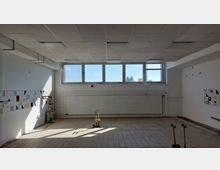 Leerer Industrie- oder Funktionsraum mit gefliesten Böden und Wänden; große rechteckige Fenster an der Stirnseite lassen Tageslicht herein, flankiert von weißen Kacheln mit bunten geometrischen Mustern. Im Raum befinden sich Installationsrohre und eine verschlossene Rollade an der Wand.