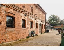 Backsteinfassade eines historischen Gebäudes mit mehreren Fenstern in Rundbogenform und dekorativen Mustern aus Ziegelsteinen; daneben ein gepflasterter Hof mit vereinzelt abgestellten Gegenständen und Bäumen im Hintergrund.