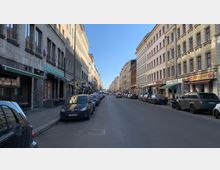 Straßenszene in einem städtischen Wohn- und Geschäftsviertel mit mehrstöckigen, historischen Gebäuden auf beiden Seiten. Entlang der Straße sind parkende Autos sowie verschiedene Geschäfte, Restaurants und eine Bäckerei sichtbar; der Himmel ist klar und blau.