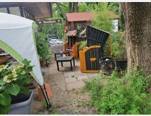 Außenbereich eines Biergartens mit Holzmöbeln und einem dunkelgeflechteten Strandkorb. Pflanzen, ein großes Zelt und ein Baum umgeben die gepflasterte Fläche, im Hintergrund ist ein Schild mit der Aufschrift „Rodelhütte“ sichtbar.