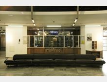 Ein Eingangsbereich mit einer breiten schwarzen Ledercouch im Vordergrund und einer Kasse für ein Planetarium im Hintergrund. Der Bereich ist schlicht gestaltet, mit weißen Wänden, gläsernen Fenstern an der Kasse und einer Neonaufschrift „KASSE PLANETARIUM“.