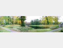 Ein weitläufiger Park mit einem kleinen See in der Mitte, umgeben von Bäumen mit herbstlich gefärbtem Laub. Rechts im Bild eine steinerne Brücke und ein gepflasterter Weg, der sich entlang des Ufers schlängelt.