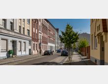 Straßenszene mit Altbau-Reihenhäusern in verschiedenen Pastellfarben, gepflastertem Bürgersteig und jungen Bäumen entlang der Straße; parkende Autos sind am Fahrbahnrand zu sehen, und der Himmel ist klar und blau.