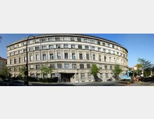 Großes historisches Gebäude mit einer geschwungenen Fassade, mehreren Stockwerken und vielen rechteckigen Fenstern; die Fassade ist aus hellem Stein. Vor dem Gebäude stehen Bäume und ein Baukran, es gibt auch parkende Autos und Straßenlaternen entlang der Straße.