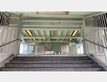 Treppe in einem U-Bahnhof mit hellgrüner Wandverkleidung und Deckenstützen. An der Wand befinden sich Schilder mit dem Stationsnamen „Warschauer Str.“, eine Informationskarte, Graffiti sowie eine verschlossene Tür.