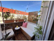 Kleiner Balkon mit Holzboden und gemütlicher Sitzecke aus weiß gepolsterten Möbeln, dekoriert mit Pflanzen in Töpfen und einer Rankpflanze an der Wand. Im Hintergrund ist ein Wohngebäude mit roten Ziegeldächern und blauen Himmel zu sehen.