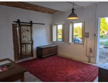 Ein rustikaler Raum mit weißen Wänden und einer hölzernen Schiebetür auf einer Ziegelwand. Der Raum ist mit drei Fenstern ausgestattet, die Aussicht auf eine grüne Landschaft bieten, ein roter Teppich bedeckt den Boden, und eine Hängelampe beleuchtet die Szene.