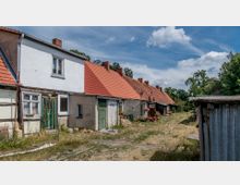 Reihenhäuser mit roten Ziegeldächern und verputzten Fassaden, teils mit sichtbaren Fachwerkelementen, in einer ländlichen Umgebung; ein unbefestigter Boden und mehrere Blumenarrangements prägen den Vordergrund, umgeben von Bäumen und Gebüsch.