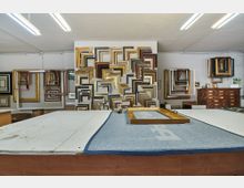 Werkstatt für Bilderrahmen mit einer großen Auswahl an unterschiedlich geformten und verzierten Rahmen an den Wänden. Im Vordergrund ein großer Holztisch mit einem blauen Tuch und einem Rahmen darauf, umgeben von Werkzeugen.