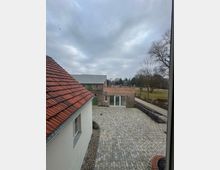 Blick auf einen gepflasterten Innenhof, umgeben von einem weiß verputzten Gebäude mit roten Dachziegeln und einem Backsteingebäude mit großen Fenstern. Im Hintergrund sind Bäume und eine Wiese unter bewölktem Himmel zu sehen.