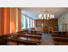 Ein kleiner Versammlungsraum mit Holzbänken und Tischen, einer hölzernen Rückwand mit integrierter Empfangstheke und modernen Hängeleuchten. Große Fenster mit orangefarbenen Vorhängen lassen Tageslicht herein, und die Decke besteht aus einem hellen Rastermuster.