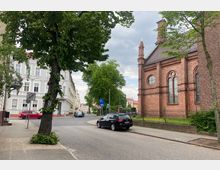 Straßenszene in einer kleinen Stadt mit einem Gehweg, parkenden Autos und einem großen Baum im Vordergrund. Rechts im Bild steht eine rote Backsteinkirche mit Rosettenfenstern und Spitzbögen, während links helle Wohngebäude mit traditionellen Fassaden zu sehen sind.