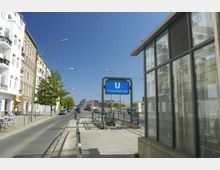 Straßenszene mit einem Eingang zur U-Bahn-Haltestelle „Schwartzkopffstraße“, erkennbar an einem blauen U-Bahn-Schild. Im Hintergrund sind Wohnhäuser, Straßenlaternen, geparkte Autos und ein Gehweg entlang der Straße zu sehen, mit klarem blauem Himmel darüber.