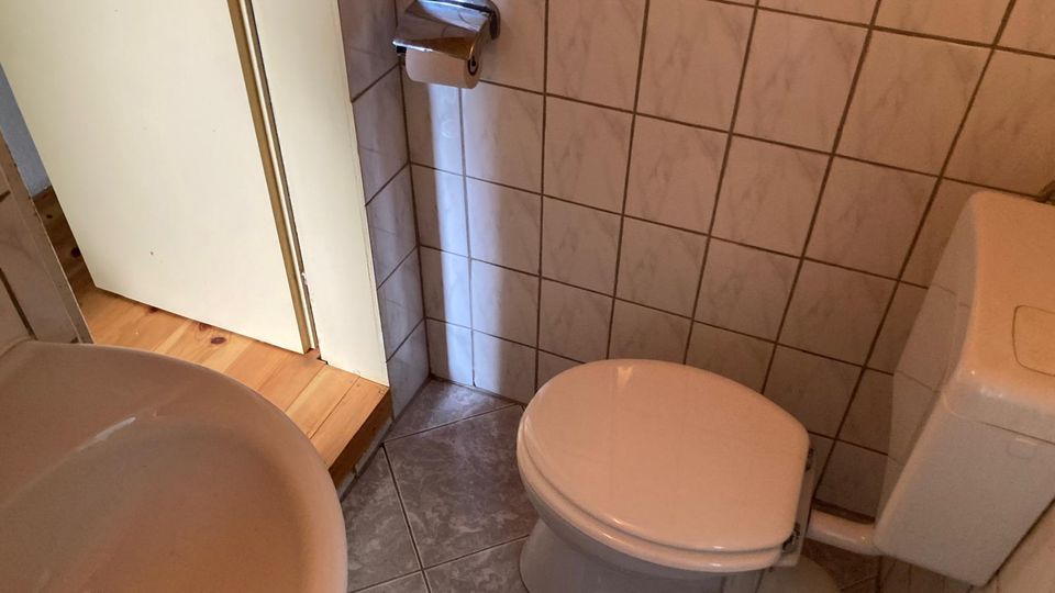 Kleines Badezimmer mit gefliesten Wänden und grauem Fliesenboden, mit WC und Wandspülkasten sowie einem Waschbecken im Vordergrund. An der Wand neben der Tür befindet sich ein Toilettenpapierhalter.