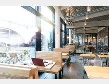 Innenansicht eines modernen Cafés mit großen Glasfronten, die auf eine Terrasse mit Tischen und Stühlen blicken. Holzsitzbänke und Tische im minimalistischen Design sind mit dezenten Dekorationen bestückt, daneben warme Beleuchtung mit freihängenden Glühbirnen und ein industriell gestaltetes Deckenkonzept.