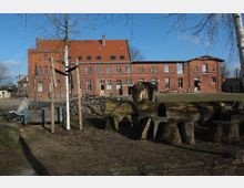 Backsteinbau mit rotem Ziegeldach in ländlicher Umgebung, mit großer Rasenfläche im Vordergrund. Vor dem Gebäude stehen Holzsitzgruppen und Baumstämme, umgeben von kahlen Bäumen und rustikaler Landschaft.