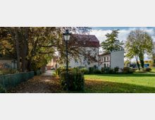 Ein gepflasterter Weg, gesäumt von Bäumen und einem grünen Zaun, führt zu einem hellen, historischen Gebäude mit rotem Satteldach. Neben dem Gebäude befindet sich eine Wiese mit einzelnen Bäumen, und im Hintergrund sind geparkte Busse erkennbar.