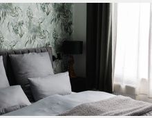 Ein modernes Schlafzimmer mit grauem gepolstertem Bett, hellblauer Bettwäsche und grauer Decke. Die Wand ist mit tropischer Blättertapete gestaltet, daneben steht eine grüne Tischlampe auf einem dunklen Nachttisch, und rechts befindet sich ein Fenster mit halbtransparentem Vorhang.