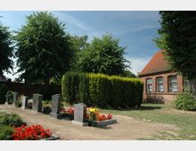 Ein kleiner Friedhof mit mehreren Grabsteinen, die von Blumenbeeten umgeben sind, im Vordergrund; im Hintergrund stehen Bäume und eine Hecke sowie ein Backsteingebäude mit roten Ziegeldach und weißen Fenstervorhängen.
