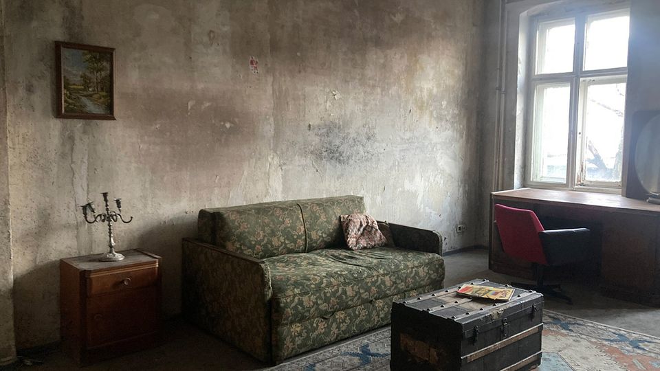 Abgenutztes Wohnzimmer mit stark verschmutzten, unverputzten Wänden, einem gemusterten Sofa und einem kleinen Beistelltisch mit Kerzenleuchter. Neben einem großen Fenster stehen ein Schreibtisch mit Stuhl; auf einem Teppich liegt eine große Holztruhe als Couchtisch.