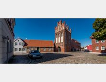 Historisches Stadtzentrum mit einem Backsteintorhaus im gotischen Stil, flankiert von niedrigen Fachwerkhäusern mit roten Ziegeldächern. Der gepflasterte Platz ist von parkenden Autos gesäumt, im Hintergrund blauer Himmel und weitere Gebäude.
