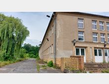 Ein zweistöckiges, rechteckiges Gebäude mit verputzter Fassade und zahlreichen weißen Fenstern; der Eingangsbereich ist mit gelben Ziegeln eingefasst. Im Vordergrund ist eine asphaltierte Straße zu sehen, rechts eine Treppe, und links befinden sich Bäume, darunter eine große Trauerweide.