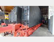 Rückansicht eines historischen schwarzen Lokomotiv-Tenders mit roten Fahrgestellen, fotografiert in einer Halle mit Holzdecke und gefliesten Wänden. Im Hintergrund ist ein gelbes Fahrzeug teilweise sichtbar.