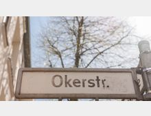Straßenschild mit der Aufschrift „Okerstr.“ vor einem Hintergrund aus einem Baum mit kahlen Ästen und einem Gebäudefassadenausschnitt bei sonnigem Wetter.