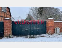 Schneebedecktes Eingangstor aus dunklem Holz mit roten Buchstaben, die das Wort „WILLKOMMEN“ formen, eingerahmt von roten Backsteinpfeilern; im Hintergrund ein Fachwerkhaus und winterliche Bäume.