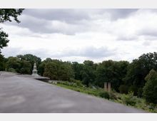 Blick auf eine bewaldete Parklandschaft mit einem kleinen mehrstöckigen Turm mit goldener Spitze, der zwischen den Bäumen hervorschaut. Im Vordergrund ist ein steinerner Geländerabschnitt zu sehen, im Hintergrund erstreckt sich dichter Wald unter einem bewölkten Himmel.