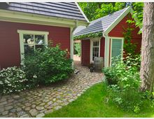 Zwei kleine rote Holzhäuser mit weißen Fensterrahmen und schwarzen Ziegeldächern, verbunden durch einen gepflasterten Weg. Umgeben von grüner Vegetation, Blumenbeeten und einem Baumstamm im Vordergrund.