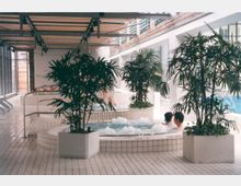 Innenbereich eines Schwimmbades mit weiß gefliesten Böden und Wänden, mehreren Whirlpools, in denen sich Menschen entspannen, und großen, grünen Pflanzen in weißen Pflanzkübeln. Im Hintergrund sind Glaswände und ein Schwimmbecken sichtbar.