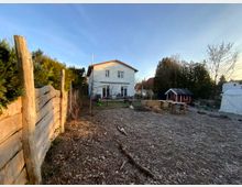 Wohnhaus mit weißer Fassade in einem großen Garten mit Baumbestand. Im Vordergrund ein rustikaler Holzzaun links, ein Bereich mit Holzschnitzeln und einfachem Holzspielgerät rechts sowie ein kleines rotes Gartenhäuschen mit Solarpanel.