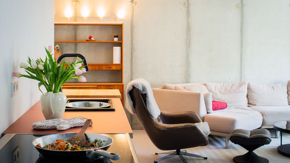 Offener Wohn- und Küchenbereich mit Betonwänden, einer Kücheninsel mit Kochfeld und Pfanne sowie einem Regal mit Beleuchtung im Hintergrund. Daneben stehen ein Ecksofa, ein Leder-Loungesessel mit Hocker und ein niedriger Couchtisch auf einem Teppich.