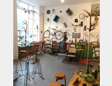 Ein kleiner Laden oder Atelier mit einer Mischung aus Vintage-Dekorationen und Möbeln. An den Wänden hängen ein Fahrrad, Metallobjekte und andere Kunstwerke, während im Raum Stühle, Glastische, stilisierte Hirschtrophäen und Vitrinen mit verschiedenen Gegenständen zu sehen sind.